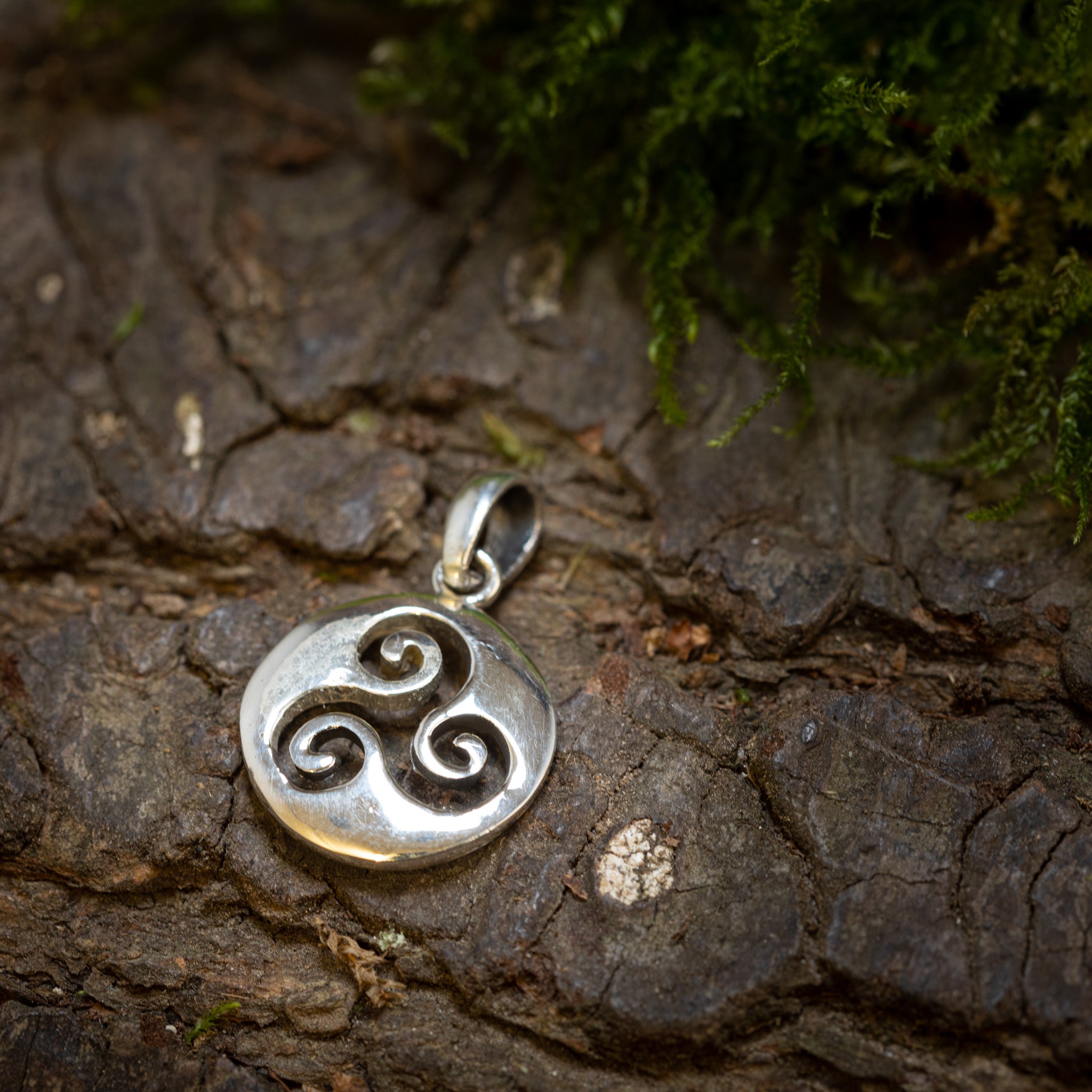 På en struktureret træbark-overflade ligger Vedhæng Åben Triscele 925s Sterling Sølv-vedhæng elegant med et triskelion-symbol på tre indbyrdes forbundne spiraler. Designet er lavet af ægte sølv, og designet er smukt indviklet, med grønt mos, der delvist dækker barken over det i dette nærbillede, der fremhæver dets udsøgte håndværk.