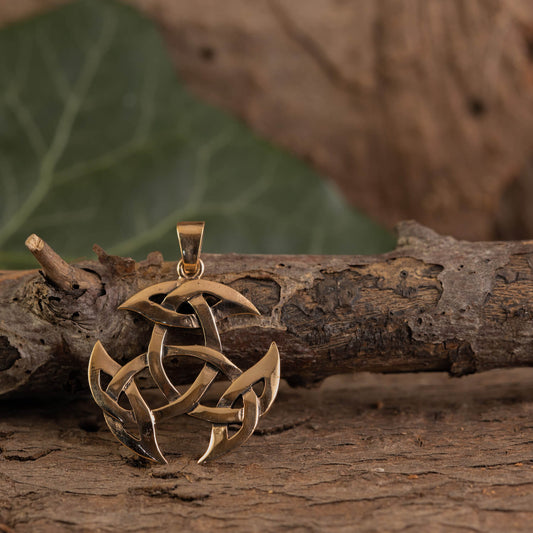 En bronze Kosmisk Hjul Vedhæng, indviklet designet med tre sammenlåsende løkker som et guld triquetra vedhæng eller middelaldersmykke, hviler på et stykke groft, tekstureret træ. Baggrunden har et sløret grønt blad og flere træteksturer.