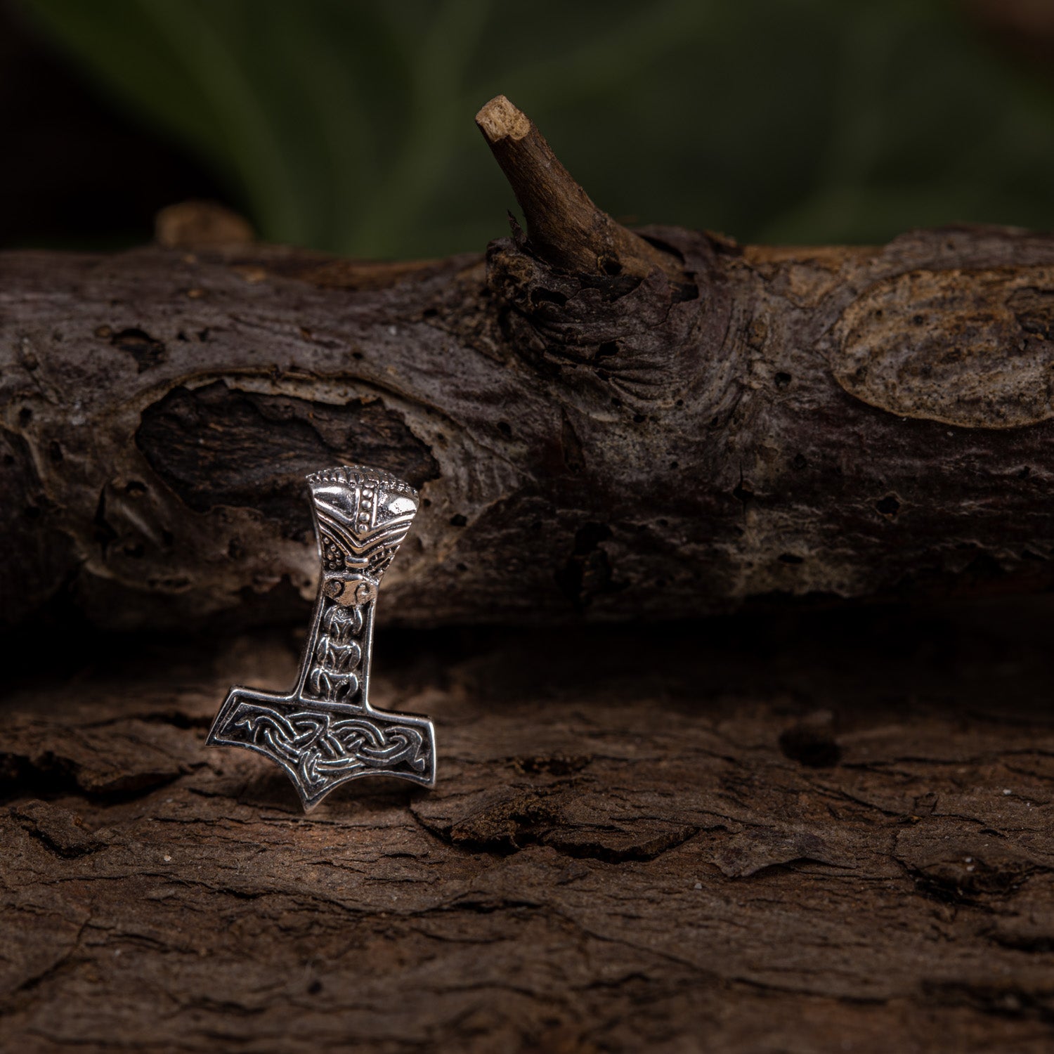 En detaljeret Thors Hammer Vedhæng 925s Sølv Mini, formet som Thors hammer og lavet af ægte sterling sølv, prydet med indviklede nordiske designs, hviler mod en tekstureret trægren. Træbaggrunden står i kontrast til vedhængets metalliske glans og fremhæver dens håndværk i en diskret vikingesmykke.