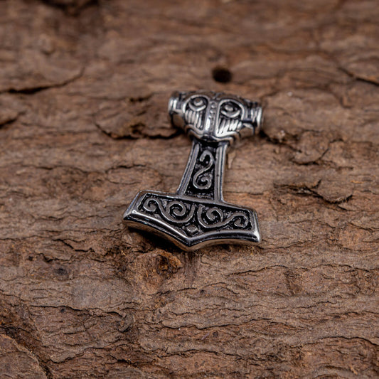 Et Thors Hammer Vedhæng Stål-vedhæng, formet som Mjölnir med indviklede nordiske designs og mønstre, hviler på en ru, tekstureret træoverflade. Dette værk afspejler vikingetidens kunstnerskab og hylder nordiske mytologi.