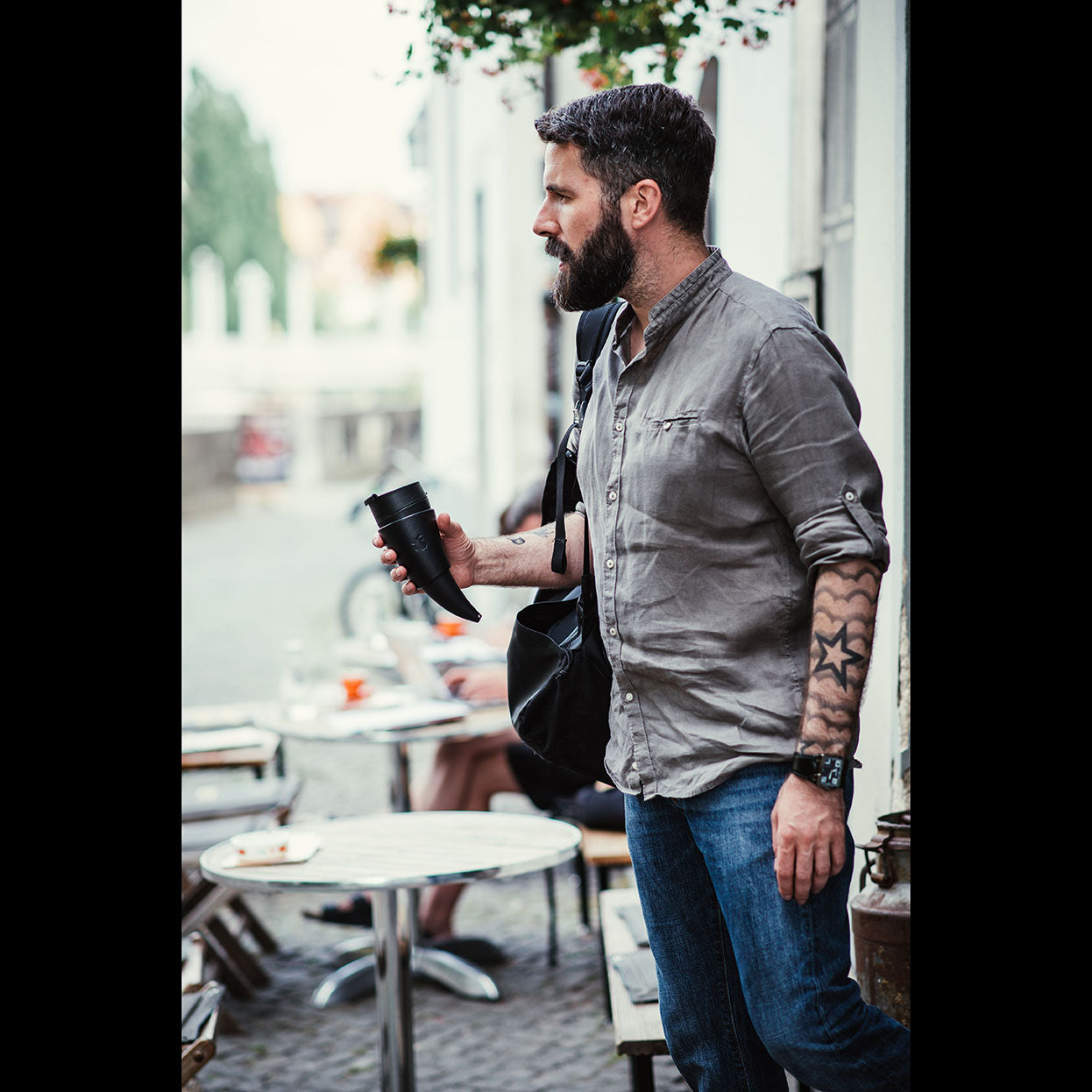 En skægget mand iført en grå skjorte og jeans står udenfor en café med en Gede Kaffe Drikkehorn Sort. Han bærer en sort skuldertaske og har et tatoveringsærme på venstre arm. Der er tomme borde og stole omkring ham, med slørede individer i baggrunden.