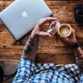 Indlæs billede i Gallery viewer, En person med tatoveringer på underarmen, iført en ternet skjorte, sidder ved et træbord. De holder en kop med latte art i den ene hånd og åbner låget på deres Ged Kaffe Drikkehorn Brun med den anden. En bærbar computer og et kamera er også på bordet, og fuldender scenen for denne moderne viking.
