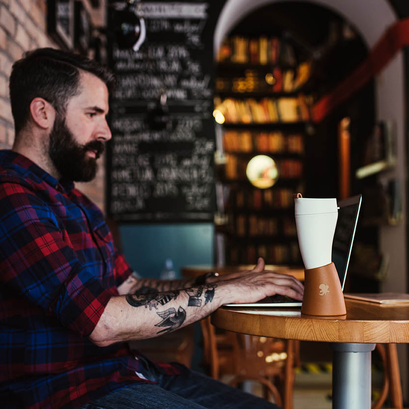 En skægget mand med tatoveringer, der ligner en moderne viking, er iført en rød og blå plaid skjorte, mens han taster på en bærbar computer ved et rundt træbord i en hyggelig café med bogreoler og tavlevægge. En ged Kaffe Drikkehorn Brun isoleret kop lavet af BPA-fri plastik sidder tæt på hans bærbare computer.