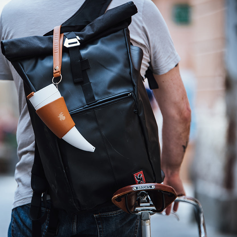 En person iført en grå t-shirt bærer en stor sort roll-top rygsæk med en hvid og solbrun ged Kaffe Drikkehorn Brun påsat, mens han skubber en cykel med en brun Brooks-sadel i læder. Scenen fremkalder stemningen fra en moderne viking på en bygade.