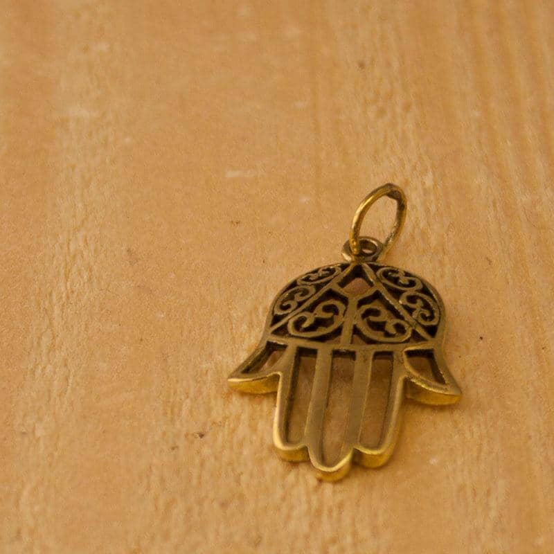 Et nærbillede af et Hamsa Vedhæng Messing vedhæng liggende på en træoverflade. Bronzevedhænget har indviklede filigrandetaljer i toppen, mens resten af hånden har et enkelt, usminket design. Træet har en let, naturlig finish.