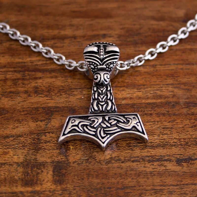 Et nærbillede af en sølvhalskæde med et vedhæng formet som Thors hammer (Mjölnir). Vedhænget har indviklede, indgraverede detaljer med nordisk inspireret design og er fastgjort til en robust Halskæde Ankerkæde i Stål 5mm. Den hviler elegant på en træoverflade.