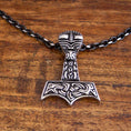 Indlæs billede i Gallery viewer, Et detaljeret vedhæng i form af Thors hammer, Mjölnir, hænger fra en Halskæde Ankerkæde Sort Stål 5mm. Vedhænget har indviklede, nordisk-inspirerede udskæringer og mønstre i en slående kontrastfarve. Den hviler på en træoverflade.
